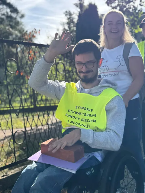 Uczestnik sztafety na wózku w żółtej kamizelce Bytomskiego Stowarzyszenia, z cegiełką na kolanach, macha do zdjęcia.