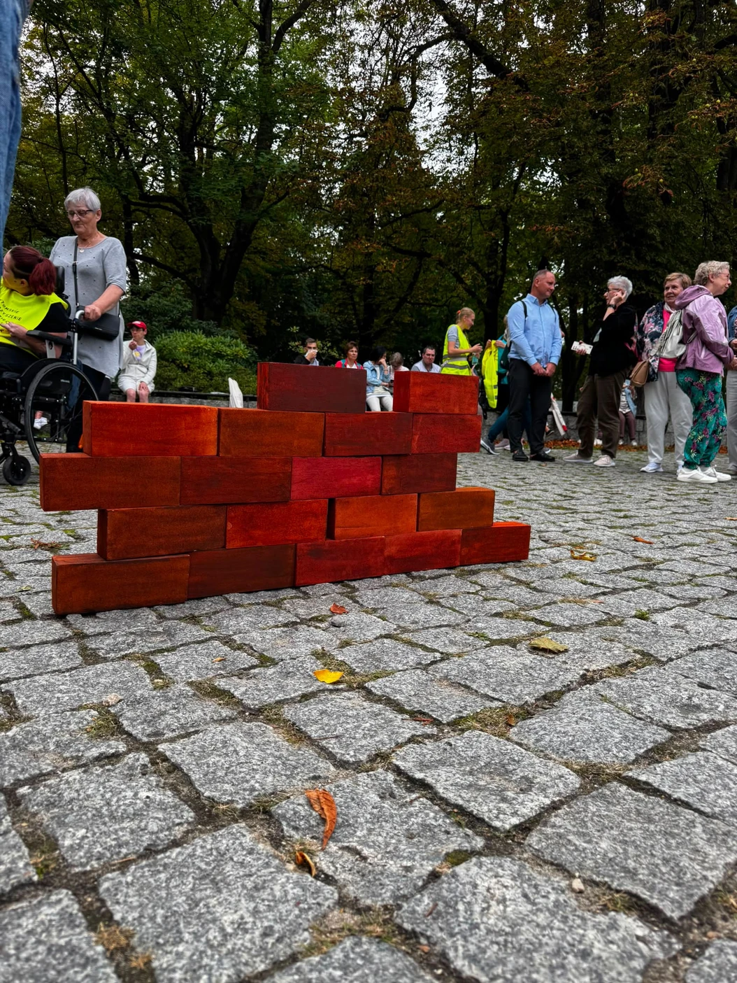Symboliczne cegiełki ułożone w ścianę na kostce w parku, w tle uczestnicy i kibice sztafety.