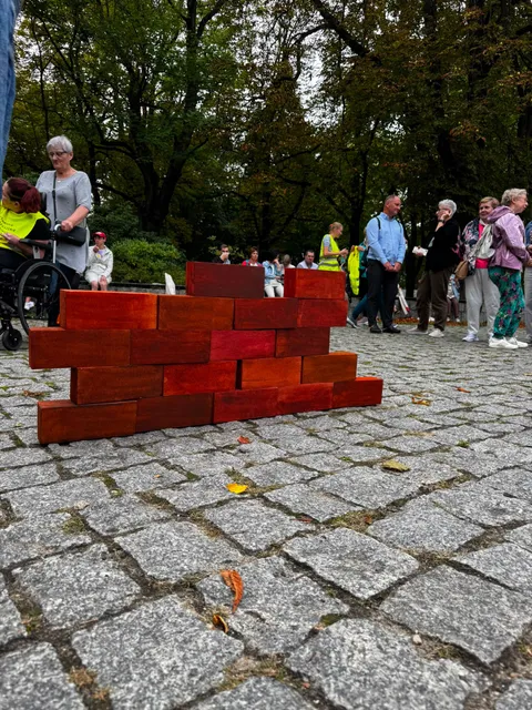 Symboliczne cegiełki ułożone w ścianę na kostce w parku, w tle uczestnicy i kibice sztafety.