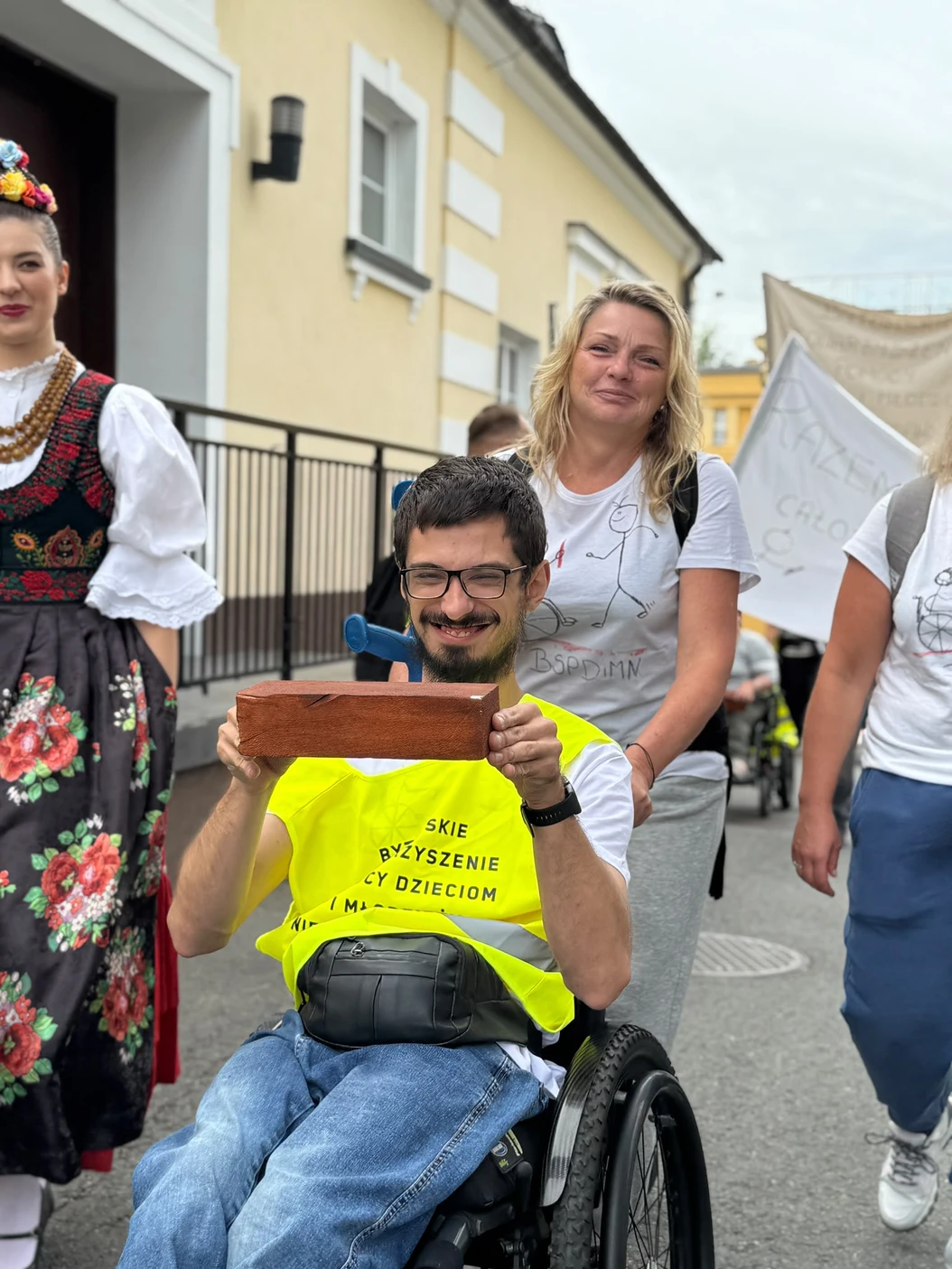 Uczestnik sztafety na wózku trzyma symboliczną cegiełkę; obok osoba w stroju ludowym i opiekunowie.