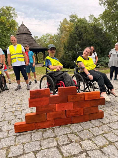 Uczestnicy sztafety przy ułożonej ze symbolicznych cegiełek ścianie w parku.