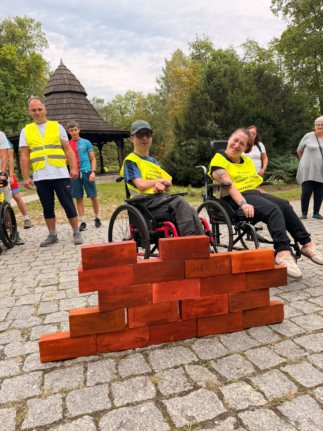 Uczestnicy sztafety przy ułożonej ze symbolicznych cegiełek ścianie w parku.
